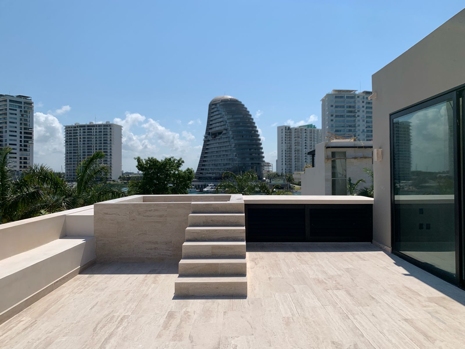 Roof garden de Residencia Lote 20 en travertino con jacuzzi elevado, escalones y vista a las torres y la marina de Puerto Cancún