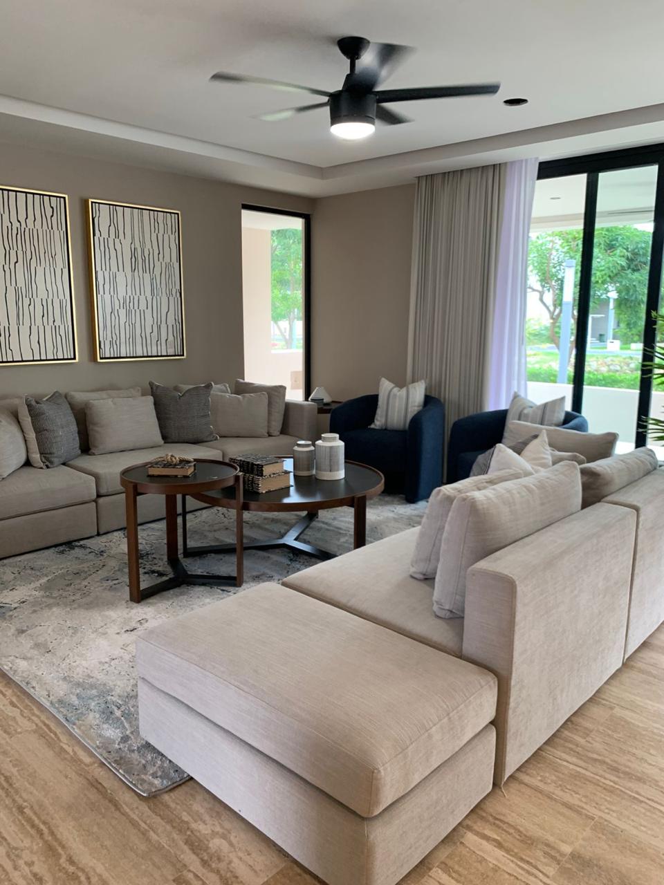 Sala de Residencia Lote 20 con sofás en tonos arena, mesas de centro redondas y ventanales al jardín